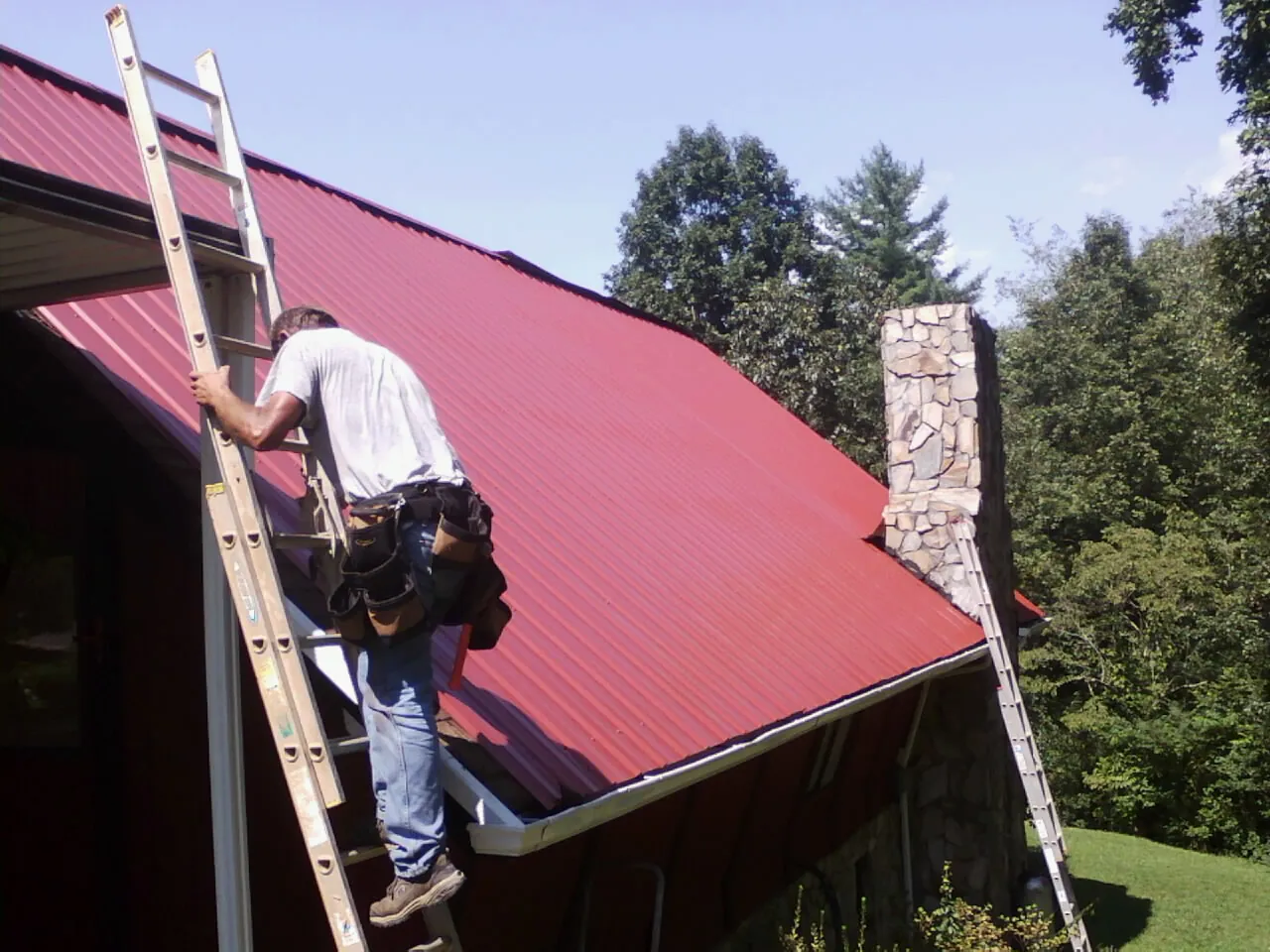 Roofer on a ladder beside a red metal roof and stone chimney — Soffit & Fascia Repair by Roofing Brothers in Barnett